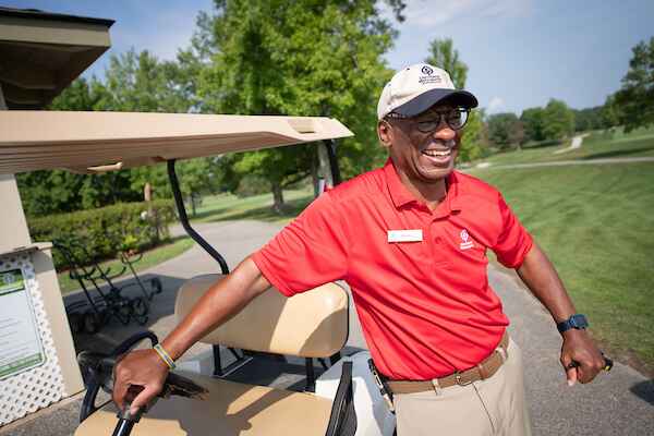 A man smiling while standing next to a golf cart on a sunny day.