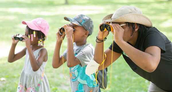 A woman wearing a safari hat and two young children looking through binoculars at something in the distance. 