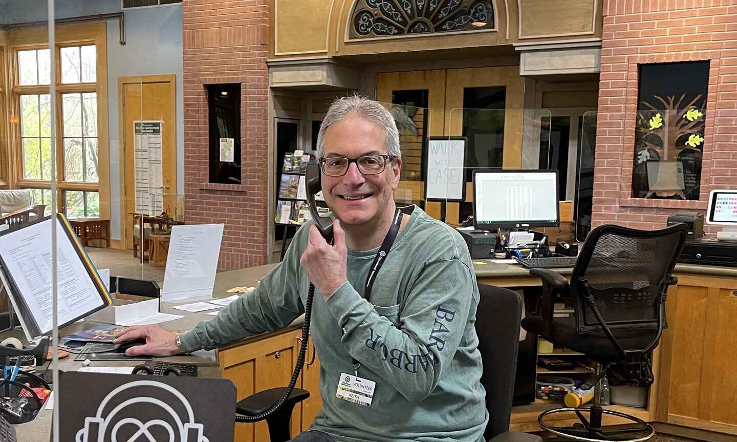 A man sitting at an information desk in front of a computer, holding a phone, and smiling.