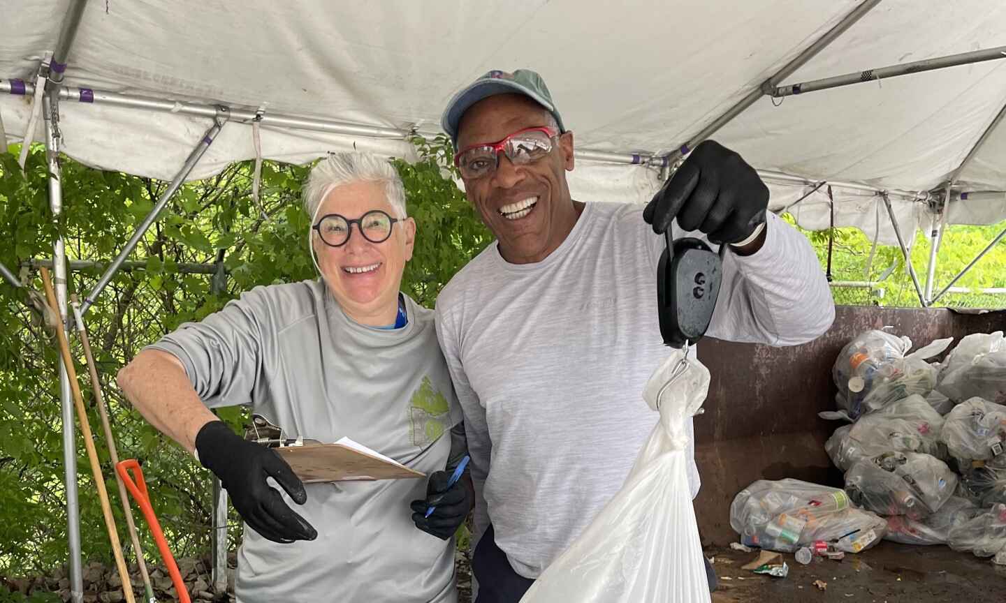 Two smiling individuals wearing gloves weigh a bag of recyclables under a tent, with bags of collected items visible in the background.