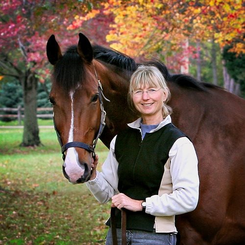 Woman standing beside a brown horse, smiling, with colorful autumn trees in the background.